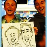 Two smiling guys proudly holding their caricature drawing by artist Grant at an event.